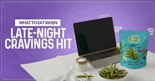 A laptop, a bag of spicy okra snacks, a bowl of dip, and scattered okra snacks are set on a table against a purple background with text: "What to eat when late-night cravings hit.